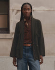 A model stands outdoors against a stone wall wearing a dark olive green open-front cardigan over a rust-red plaid shirt, paired with blue jeans and a brown belt.
