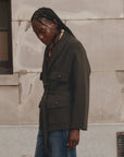 A woman wearing a dark olive green belted wrap jacket and blue jeans, standing against a textured beige stone wall.