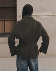 A person viewed from behind wearing an olive green belted coat and blue jeans, standing against a light beige stone wall.