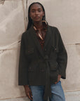 A model wearing a dark olive green knit shawl-collar jacket with a tie belt, styled over a patterned shirt and blue jeans, standing against a textured beige stone wall.
