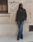A woman stands against a textured beige stone wall wearing a dark olive green belted jacket, blue wide-leg jeans, and brown shoes.