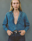 A woman modeling a cropped blue denim shirt-jacket with a deep V-neck over dark blue jeans, standing against a solid gray studio background.