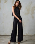 Woman wearing a black sleeveless top and wide-leg pants against a textured wall.