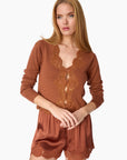A woman modeling a rust-brown long-sleeve knit top and matching satin shorts set, both featuring lace trim details, against a plain white background.