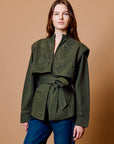 A woman modeling an olive green cropped jacket with intricate embroidery and a matching tie belt, paired with blue jeans, against a solid tan studio background.