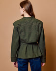 A woman viewed from the back wearing an olive green cropped jacket with decorative cutout embroidery on the back, paired with blue jeans, against a solid tan studio background.