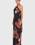 A woman modeling a black halter-neck maxi dress with a vibrant red, orange, and white paisley and floral print, set against a solid light gray background.