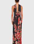 A woman viewed from the back wearing a sleeveless, floor-length halter dress with a bold floral print in red, orange, cream, and black on a light gray studio background.