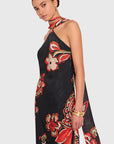 A woman modeling a black halter-neck maxi dress with a vibrant red, orange, and beige floral print, set against a solid light gray background.