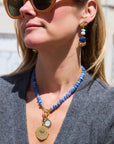 A woman wearing a blue beaded necklace with gold-tone charms and matching drop earrings, photographed outdoors against a blurred light-colored building background.