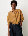 A model wearing a mustard yellow button-down shirt with thin vertical stripes and dark blue high-waisted wide-leg jeans, standing against a plain off-white background.