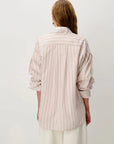 A woman shown from the back wearing a light pink and beige vertically striped button-up shirt with rolled-up sleeves, paired with white trousers, against a plain light gray background.