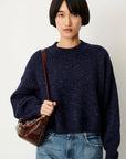 A model with a short dark bob haircut wearing an oversized navy blue heathered knit sweater and blue jeans, carrying a brown leather shoulder bag against a plain off-white background.