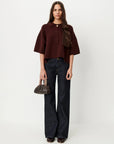 A woman stands against a plain light gray studio background wearing a cropped short-sleeve collared top in deep burgundy, dark wide-leg jeans, and holding a small dark brown handbag.