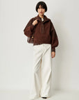 A model wearing a brown knit mock-neck sweater with textured sleeves, paired with wide-leg cream trousers and a brown shoulder bag, standing against a plain light gray background.