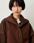 A model with a short dark bob haircut wearing a brown corduroy jacket with a high collar and button details over a patterned shirt and white undershirt, set against a plain light gray background.