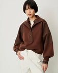 A model wearing a brown color-blocked knit pullover sweater with a high collar and white pants, photographed against a light gray studio background.