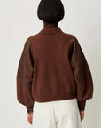 A person shown from the back wearing a brown knit sweater with darker brown patchwork sleeves and white pants, photographed against a plain light background.