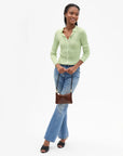 A woman modeling a light green ribbed knit button-up shirt, blue flare jeans, and black sandals, holding a small brown leather shoulder bag against a plain white studio background.