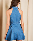 A woman wearing a blue denim halter mini dress, shown from the back against a neutral beige curtain backdrop.