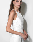 A woman modeling a white sleeveless wrap-style top and matching white bottoms against a plain white wall, with soft shadows cast on the background.