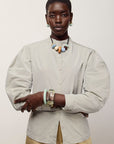 A model wearing a light beige, oversized long-sleeve button-up shirt with voluminous balloon sleeves, paired with beige trousers and colorful statement jewelry, against a plain light gray background.