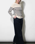 A woman modeling a cream knit sweater with black horizontal stripes on the sleeves and upper chest, paired with dark wide-leg trousers and black ankle boots, standing against a plain light gray wall.