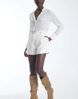 A model wearing a white long-sleeve collared shirt and matching white shorts with a gold belt, paired with tan knee-high heeled boots, against a plain white studio background.