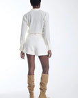 A model shown from the back wearing a white long-sleeve collared shirt, white shorts, and tan knee-high suede boots against a plain white studio background.