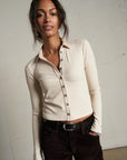 Woman wearing a beige long-sleeve shirt with buttons and dark pants against a plain background