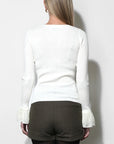 A woman shown from the back wearing a white long-sleeve knit top with ruffled cuffs, paired with dark brown shorts and black tights, against a plain white wall.