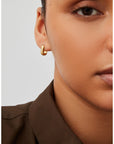 A close-up of a model wearing a small, polished gold huggie hoop earring, paired with a dark brown collared shirt against a light gray background.