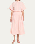 A model wearing a light pink two-piece set consisting of a short-sleeve collared blouse and a matching pleated midi skirt, paired with white heeled sandals, against a solid off-white background.