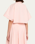 A model shown from the back wearing a light pink two-piece outfit consisting of a short-sleeved cropped top with a collar and a matching pleated skirt, against a plain off-white background.