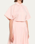 A model wearing a light pink two-piece set consisting of a short-sleeve collared blouse and a matching pleated skirt, shot against a solid off-white background.