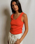 Woman wearing an orange tank top and white pants against a plain background
