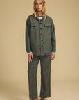 A woman modeling a matching two-piece olive green set consisting of a long-sleeve button-front shirt jacket and wide-leg trousers, styled with a light blue striped shirt and brown slip-on shoes against a solid beige studio background.