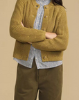 A woman modeling a mustard yellow ribbed knit cardigan over a blue and white striped shirt, paired with olive green pants, against a solid beige background.