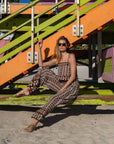 A woman wearing a brown and beige geometric patterned set sits on a colorful wooden beach staircase, with sand visible in the foreground.