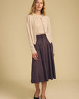 A woman modeling a light beige cardigan over a cream top and a dark purple pleated midi skirt, paired with black pointed-toe heels, against a solid beige studio background.