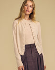 A woman modeling a light beige knit cardigan with decorative buttons over a matching beige top and a dark brown pleated skirt, set against a solid beige background.