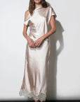 A woman models a champagne-colored satin midi dress with a draped, one-shoulder style and a lace-trimmed hem, set against a plain white wall.