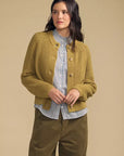 A woman modeling a mustard yellow ribbed knit cardigan with brown buttons, worn over a blue and white striped shirt and paired with olive green pants, against a solid beige background.