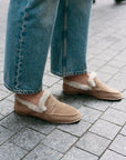 A person wearing beige suede slip-on loafers with white shearling lining, paired with blue jeans, standing on a gray paved outdoor surface.