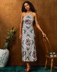 A woman models a sleeveless halter midi dress with a colorful floral and geometric diamond pattern, standing in front of a wooden wall next to a wooden chair and a potted plant.