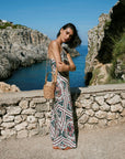 A woman stands outdoors by a rocky coastal cliffside overlooking the sea, wearing a sleeveless geometric print maxi dress and carrying a woven tan handbag.