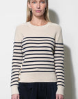 A woman modeling a cream-colored knit sweater with navy blue horizontal stripes across the chest and sleeves, paired with dark navy trousers, against a plain off-white background.