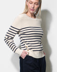 A woman wearing a cream and navy striped knit sweater with gold buttons on the front, paired with dark navy pants, standing against a light gray wall.