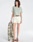Woman holding a floral jacket, wearing a light green top and white shorts on a white background