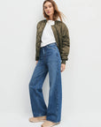 Woman wearing a green bomber jacket, white shirt, blue jeans, and beige shoes on a light gray background, side view